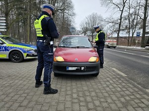 Zdjęcie przedstawia dwóch umundurowanych policjantów, oznakowany radiowóz oraz samochód osobowy.