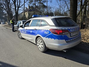 Na zdjęciu znajdują się oznakowany radiowóz, samochód osobowy oraz umundurowany policjant.