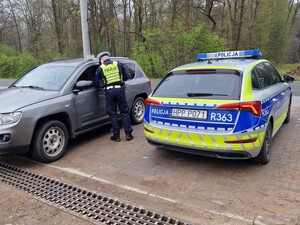 Na zdjęciu znajduje się radiowóz, samochód osobowy oraz umundurowany policjant.